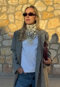 Manteau gris à chevrons par-dessus une chemise blanche, écharpe noire à motifs, et jeans bleus ; tenant un sac bordeaux et portant des lunettes de soleil foncées.