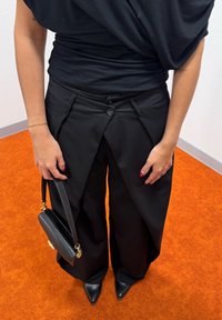 Personne portant un pantalon noir à jambes larges avec des panneaux avant uniques qui se chevauchent, un haut noir, des chaussures à bout pointu, tenant un petit sac à main noir.