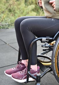 Schwarze Leggings und kastanienbraune Sportschuhe mit strukturierten, gezackten Mustern; sitzt in einem Rollstuhl mit einem schwarzen und metallischen Gestell.