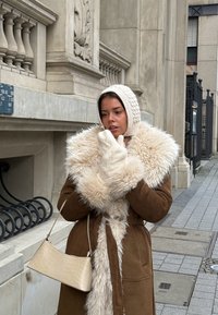 Femme ajustant une cagoule en tricot crème et des moufles, portant un manteau marron doublé de fourrure, tenant un sac à main beige, debout sur un trottoir de la ville près d'un bâtiment orné.