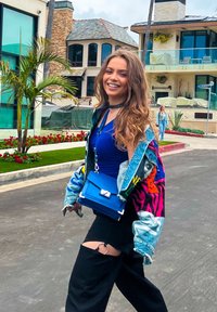 Denim jacket with colorful patterns, blue top, and black ripped pants. Holds a small blue bag with a silver clasp. Green plants in background.