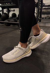 Chaussures de sport gris clair avec tissu texturé, semelles en caoutchouc et accents jaunes. Portées avec des leggings noirs ajustés dans une salle de sport.