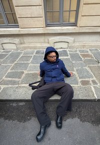 Una persona sentada en un pavimento de piedra, vistiendo una chaqueta azul con capucha, pantalones anchos negros y botas negras. Se ve una pared con ventanas detrás.