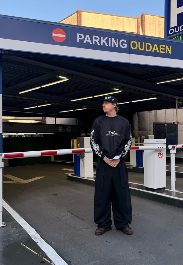 Vyras dėvi juodus platėjančius kelnes ir juodą ilgomis rankovėmis marškinius, stovintis prie uždarytos užtvaros Oudaen automobilių stovėjimo aikštelės įėjime.