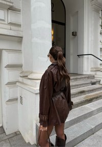 Bruine leren trenchcoat met een ceintuur in de taille, lange mouwen en knopenaccenten, gedragen over een fitted outfit. Glad textuur en casual pasvorm.