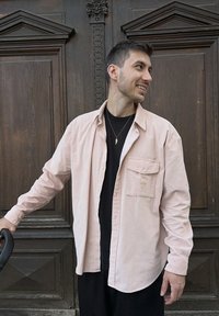 Camisa rosa claro com botões e bolso no peito, usada sobre uma camisa preta, com um fundo de madeira. Tecido liso com corte casual.