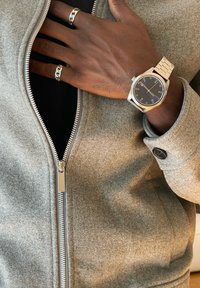 Tmavá ruka s dvěma stříbrnými prsteny a stříbrnými hodinkami spočívající na šedé bundě na zip s černým manžetem na knoflíky.