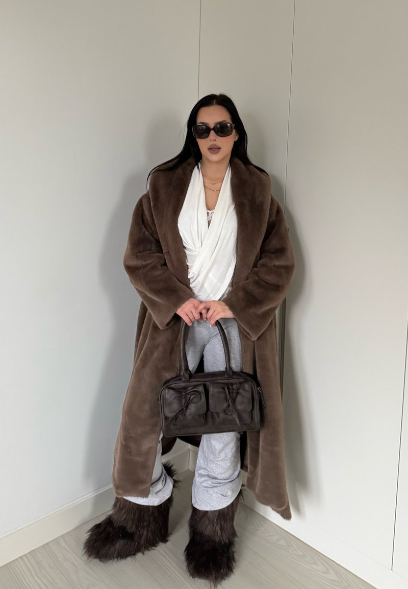 Manteau en fausse fourrure marron, haut blanc, pantalon de survêtement gris et sac marron avec détails noués. Cheveux longs noirs et lunettes de soleil foncées. Fond uni.