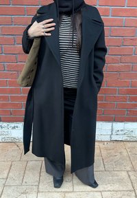 Manteau en laine noire avec un large col, porté sur une chemise rayée noire et blanche. Associé à un pantalon gris et des bottines noires. Tenant un sac vert.