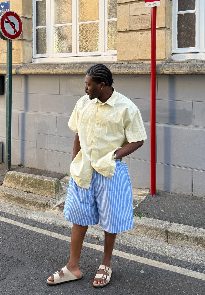 Chemise jaune clair à manches courtes, shorts rayés bleus et blancs, sandales beiges. Debout à l'extérieur près d'un panneau, avec des cheveux tressés.