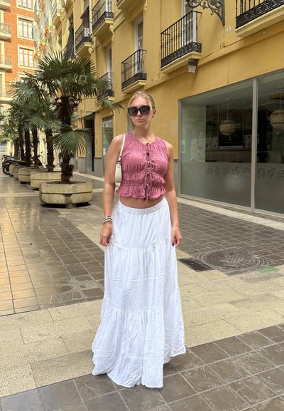 Top corto texturizado rosa con lazos frontales, combinado con una falda larga blanca y fluida. El entorno urbano presenta palmeras y pavimento de azulejos.