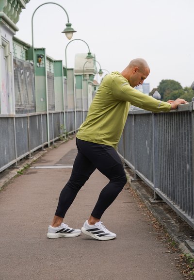 Hombre con una camiseta de manga larga verde y leggings negros estirando los músculos de las pantorrillas utilizando una barandilla metálica en una acera urbana.