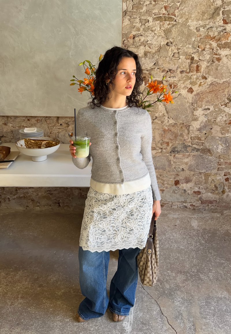 Femme en cardigan gris, jupe en dentelle portée sur un jean, tenant une boisson verte avec une paille et un sac à motifs, debout contre un mur en pierre avec des fleurs orange.
