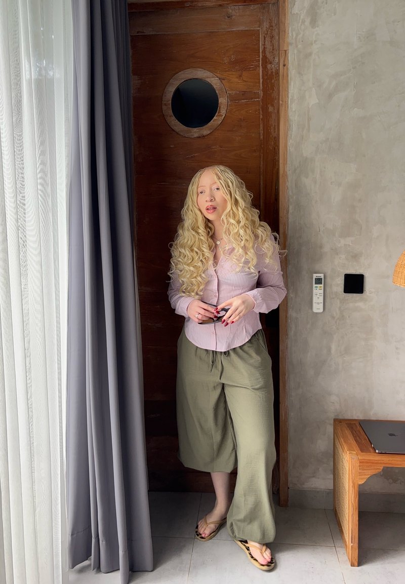 Femeie blondă cu păr creț stă lângă o ușă din lemn, purtând o cămașă liliachie deschisă, pantaloni largi de culoare măslinie, papuci de plajă, ținând un telefon într-o cameră modernă.