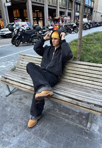 Casaco de ganga preto e calças, gorro amarelo, sabrinas bege. Sentado num banco de madeira num ambiente urbano com motocicletas estacionadas nas proximidades.