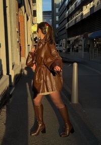 Brun skinnjakke med oversize passform og belte i midjen, kombinert med brun knelange støvler med firkantet tå. Står foran en gatebakgrunn.