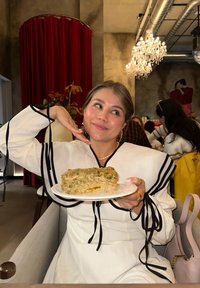 En kvinde i en hvid top med sorte kanter holder en tallerken med lagdelt lasagne. Retten har et grønt urte-topping og en cremet konsistens.