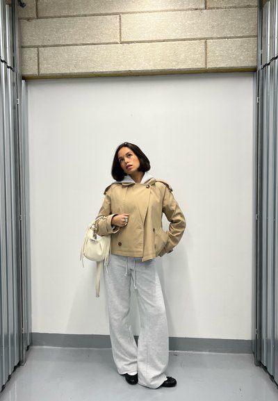 Mujer con chaqueta beige y pantalones anchos gris claro está de pie contra una pared blanca con estructuras metálicas a los lados.