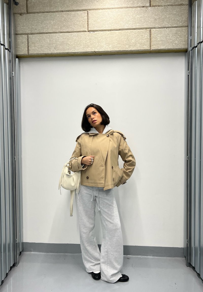 Beige dzsekit és világosszürke, bő szárú nadrágot viselő nő áll egy fehér fal előtt, melynek oldalain fém szerkezetek vannak.