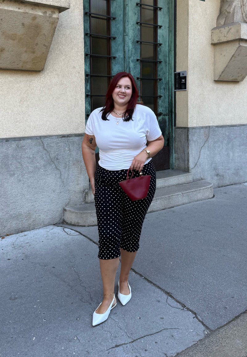 Žena s červenými vlasmi, oblečená v bielej košeli, čiernych nohaviciach s bodkami, bielych topánkach, držiaca červenú kabelku, kráča po mestskom chodníku vedľa zelenej dverí.
