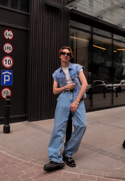 Persona joven con chaleco de mezclilla sin mangas, pantalones anchos, botas negras y gafas de sol apoyado contra un poste de la calle en una ciudad cerca de señales de tráfico.