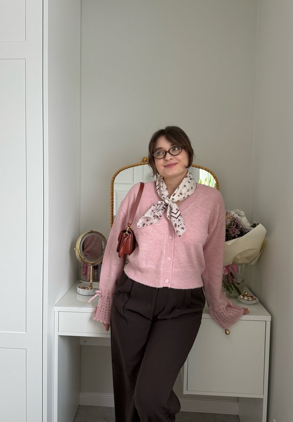 Vrouw met een bril, roze vest, bloemige sjaal en bruine broek leunt tegen een witte kaptafel met spiegel en een boeket in een lichte kamer.