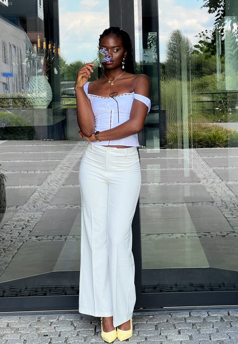 Λευκό τοπ με ανοιχτούς ώμους και ρυάκια, συνδυασμένο με φαρδιά ανοιχτόχρωμα παντελόνια και κίτρινες γόβες. Το μοντέλο κρατά ένα λουλούδι κοντά στο πρόσωπό της.