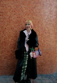 Czarny płaszcz z futrzanym kołnierzem ze sztucznego futra, na różowej bluzce, zielona spódnica w kratę. Różowa torebka z pluszowymi brelokami w kształcie lalek, okulary o okrągłych oprawkach.