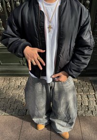 Personne portant une veste bomber noire, un t-shirt blanc, un pantalon gris ample, des bottes jaunes, un collier avec une croix en or et des bagues, faisant un geste de la main.