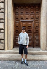 Pulover tricotat gri, pantaloni scurți negri, șosete albe și pantofi negri purtați de o persoană care stă în fața unei uși din lemn detaliate cu panouri pătrate.