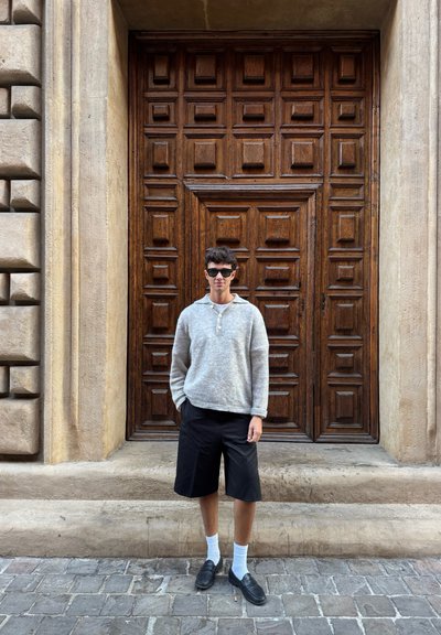 Suéter de punto gris, pantalones cortos negros, calcetines blancos y zapatos negros usados por una persona que está de pie frente a una puerta de madera detallada con paneles cuadrados.