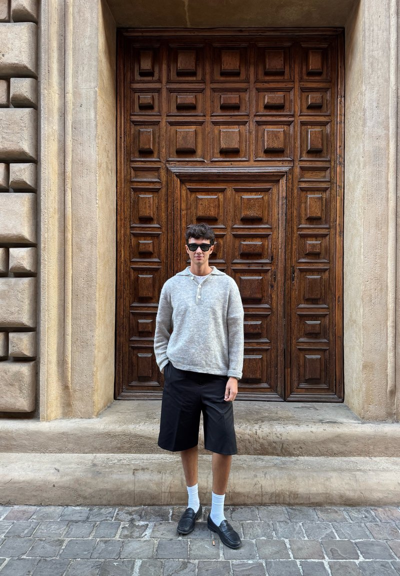 Grå stickad tröja, svarta shorts, vita strumpor och svarta skor som bärs av en person som står framför en detaljerad trädörr med fyrkantiga paneler.