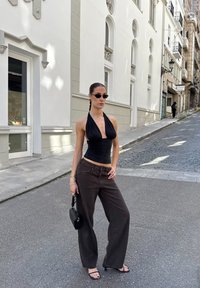 Top negro sin mangas, jeans de pierna ancha marrón oscuro y sandalias negras con tiras. Joyería de plata y un pequeño bolso negro complementan el look. Fondo de calle adoquinada.