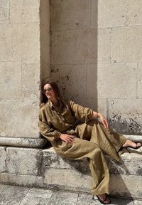Olivově zelený lněný outfit s oversize košilí a širokými kalhotami, doplněný černými sandály na vysokém podpatku, sedící na pozadí texturované kamenné zdi.