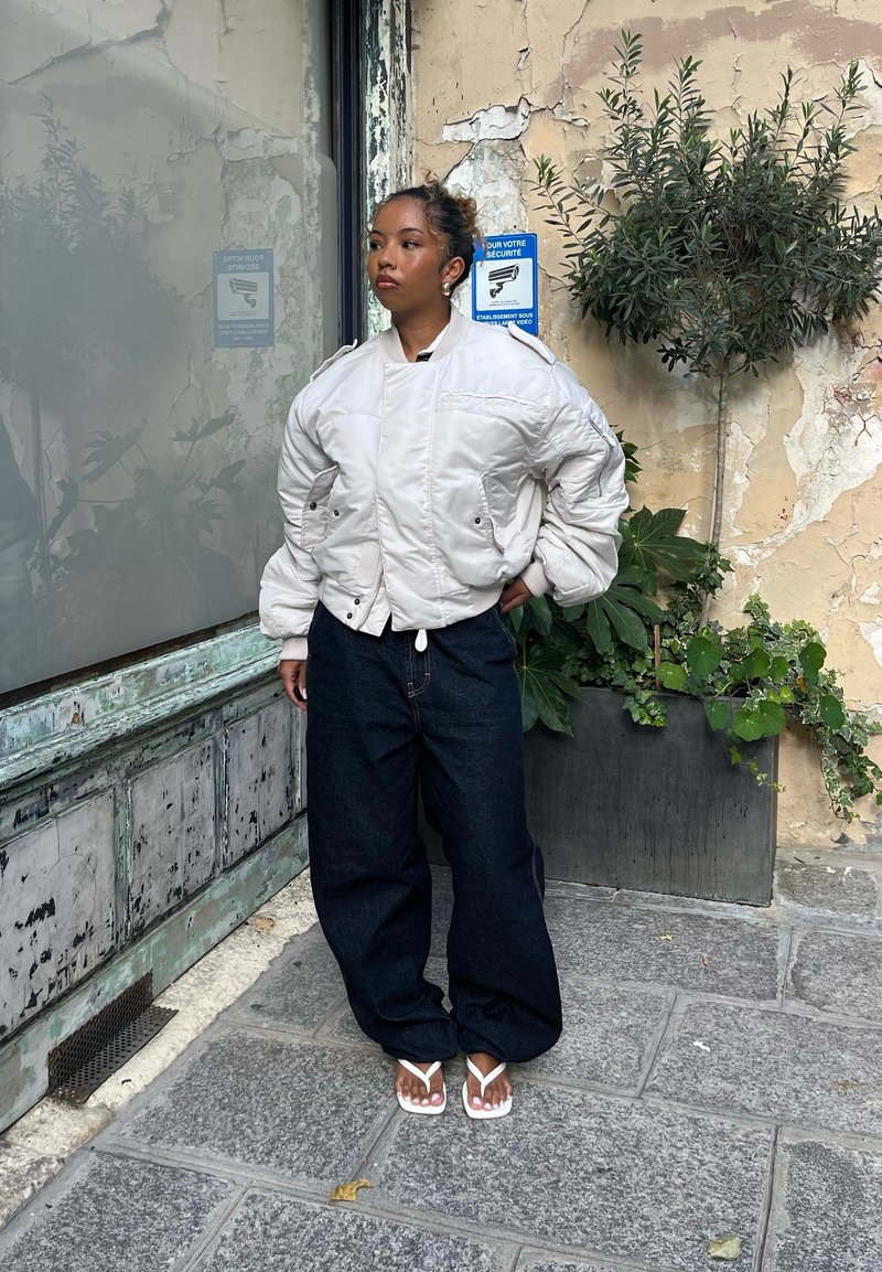 Blouson bomber blanc duveteux avec poches latérales, associé à un jean bleu large et des tongs blanches, se tient devant un mur texturé et du feuillage.