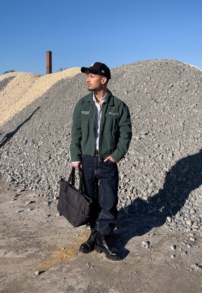 Chaqueta verde, camisa blanca, jeans azules y botas de combate negras. Sosteniendo una bolsa tote suave negra. De pie frente a montículos de grava.