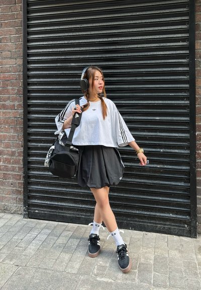 Camisa blanca corta de Adidas con rayas negras, falda negra y zapatillas negras. Bolso negro con detalles gráficos y calcetines blancos con acentos negros.