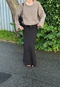 Světle hnědý pletený svetr s širokými rukávy, kombinovaný s úzkou černou maxi sukni a prošívanou černou kabelkou. Zelené listnaté pozadí.