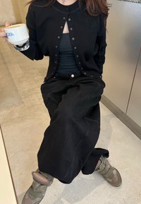 Svart cardigan med trykknapper, båret over en svart tanktop, kombinert med løse svarte jeans. Grå sko med teksturerte detaljer. Holder en hvit kopp.