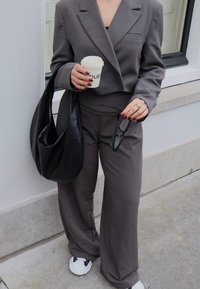 Γκρι σακάκι προσαρμοσμένο και αντίστοιχες φαρδιές παν trousers, συνδυασμένα με μια μαύρη τσάντα, άσπρα αθλητικά παπούτσια, και κρατώντας ένα φλιτζάνι καφέ.