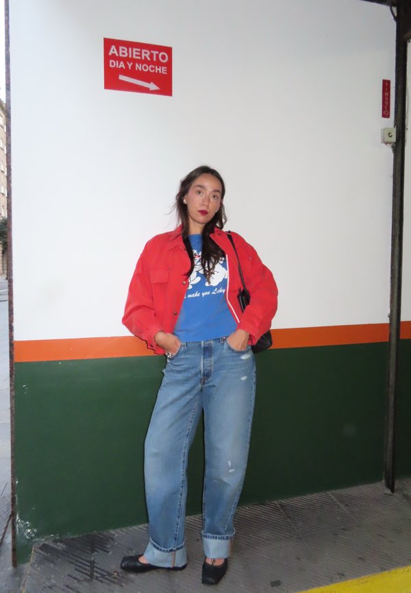 Mujer con chaqueta roja, camiseta azul con diseño gráfico y vaqueros remangados, apoyada contra una pared blanca con franjas verdes y naranjas, bajo un cartel rojo "Abierto día y noche".