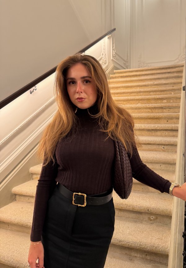 Maglia nera a costine con collo alto, gonna nera e borsa intrecciata con fibbia dorata. Sullo sfondo si vede una scala con gradini in tonalità neutra.