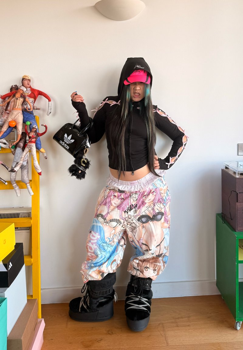 Jeune personne portant un sweat à capuche noir court, un pantalon ample imprimé anime, des bottes noires et une casquette rose, tenant un petit sac noir Adidas à l'intérieur.