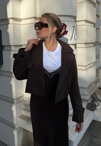 Giacca cropped marrone sopra una T-shirt bianca con bordo in pizzo, abbinata a una gonna nera. Grande occhiali da sole scuri e un accessorio per capelli color bordeaux.