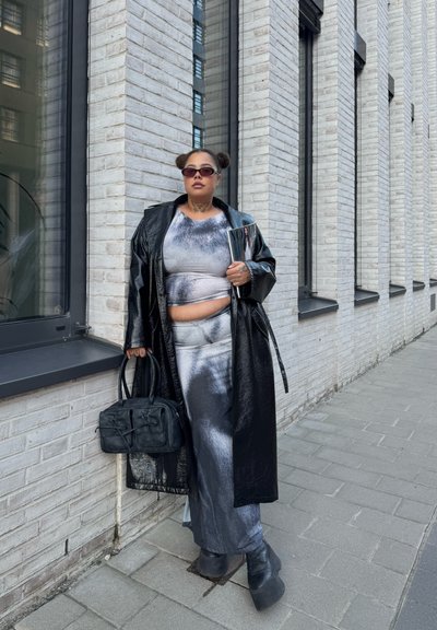 Abrigo largo negro brillante sobre un conjunto de dos piezas gris con efecto tie-dye, combinado con botas negras de plataforma y un bolso negro texturizado. Incluye pequeñas gafas de sol ahumadas.