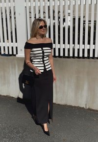 Haut à épaules dénudées rayé avec accents noirs et boutons dorés. Jupe noire longue jusqu'au sol avec une fente sur le côté. Sac à main noir et talons.