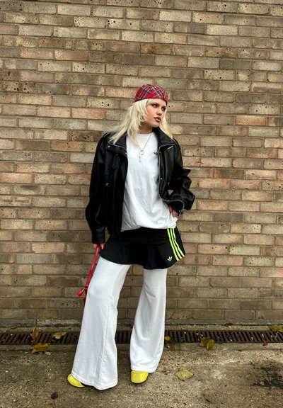 Chaqueta de cuero negra, camiseta blanca de gran tamaño, falda negra con rayas verdes, pantalones blancos holgados, zapatos amarillos, banda para el cabello a cuadros, collar plateado.