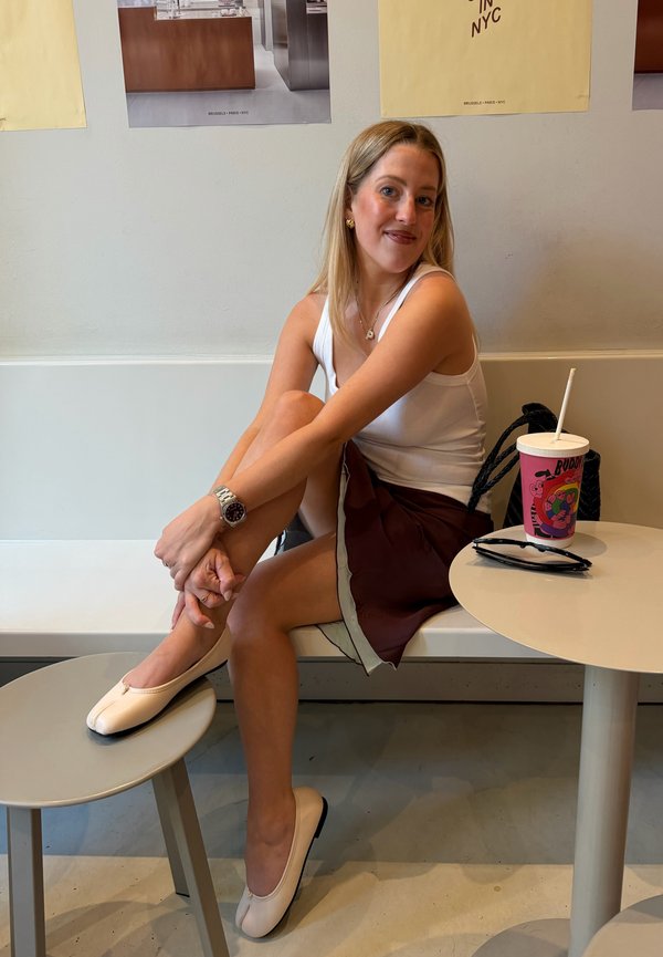 Femme en débardeur blanc et jupe marron assise sur un banc, un pied sur un tabouret, souriante, à côté d'une table avec une boisson et des lunettes de soleil.