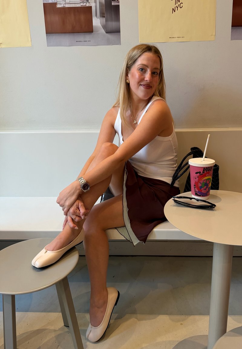 Kvinne i hvit singlet og brun skjørt sitter på en benk med en fot på en krakk, smilende, ved siden av et bord med en drink og solbriller.