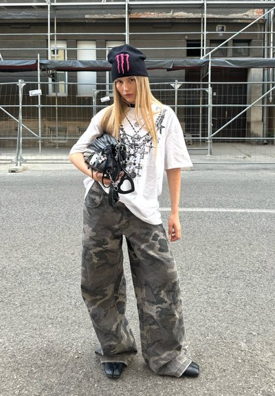 Gorro negro con logo rosa, camiseta blanca de gran tamaño con estampado gráfico, pantalones anchos de camuflaje gris y zapatos negros de punta.